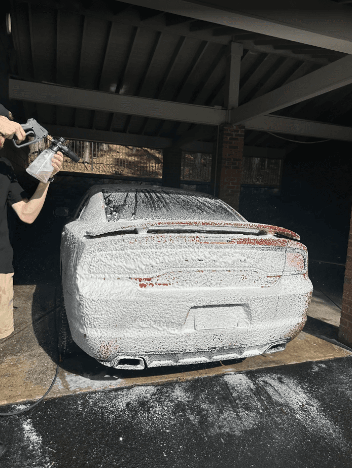 Person spraying thick white foam onto a red car parked under a carport.
