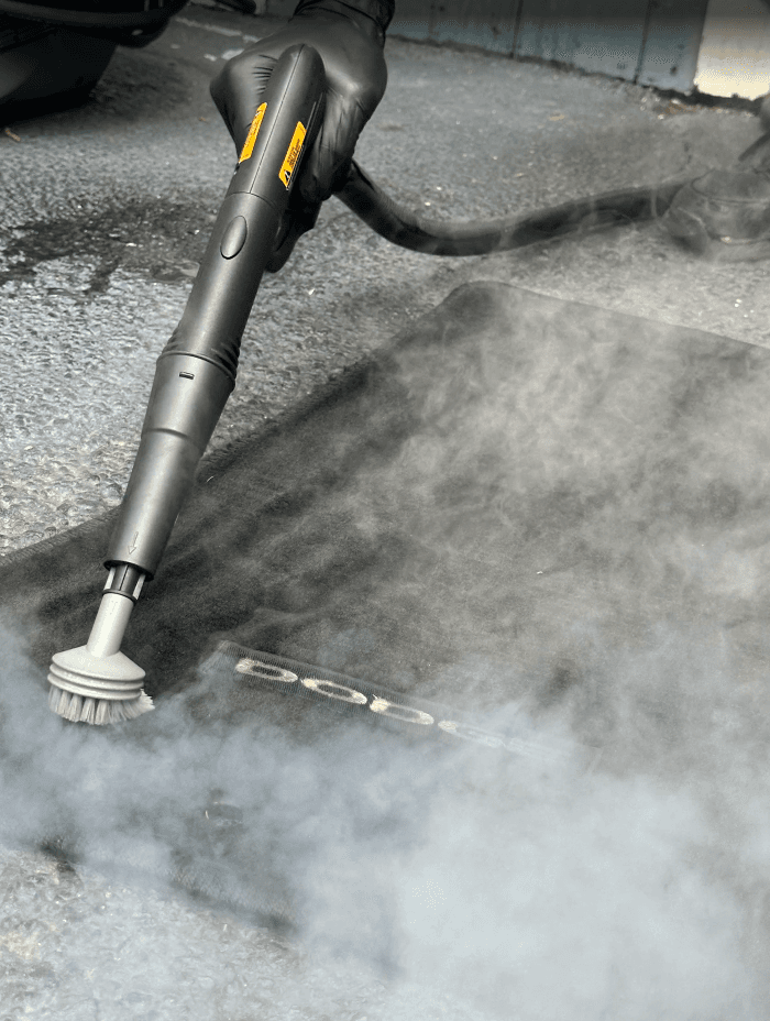 Gloved hand steam cleaning a black Dodge car floor mat with a brush tool.